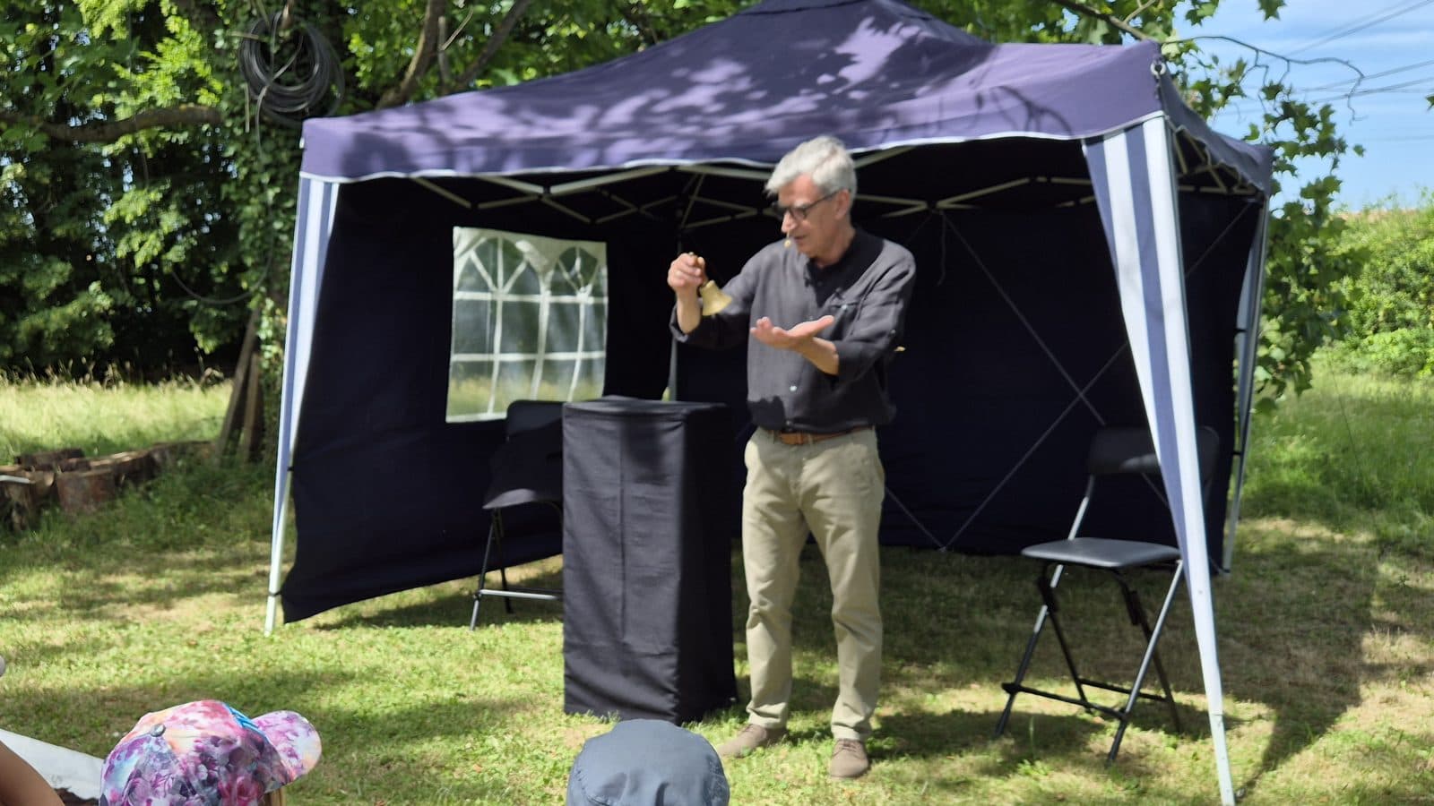 Le Magiconteur, clochette à la main, sous son barnum bleu dans le parc, devant des enfants assis dans l’herbe